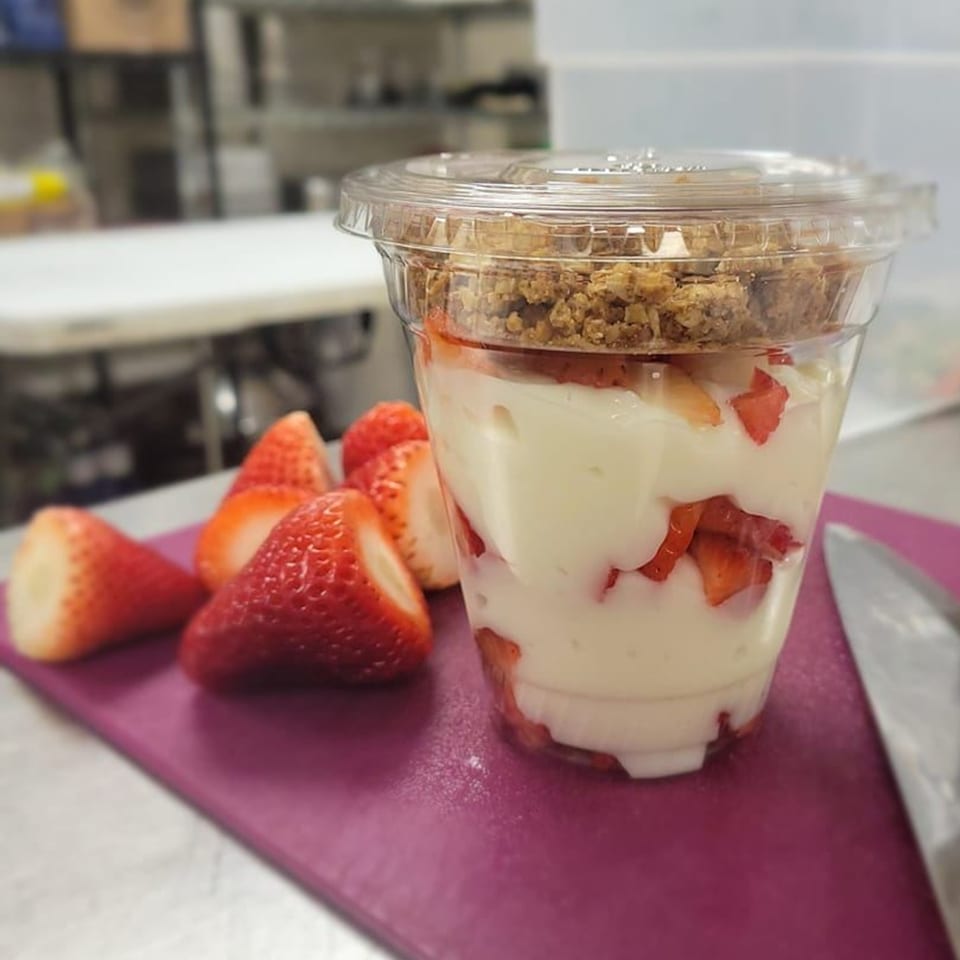 A clear plastic cup of yogurt parfait with strawberries and granola, next to fresh strawberries.