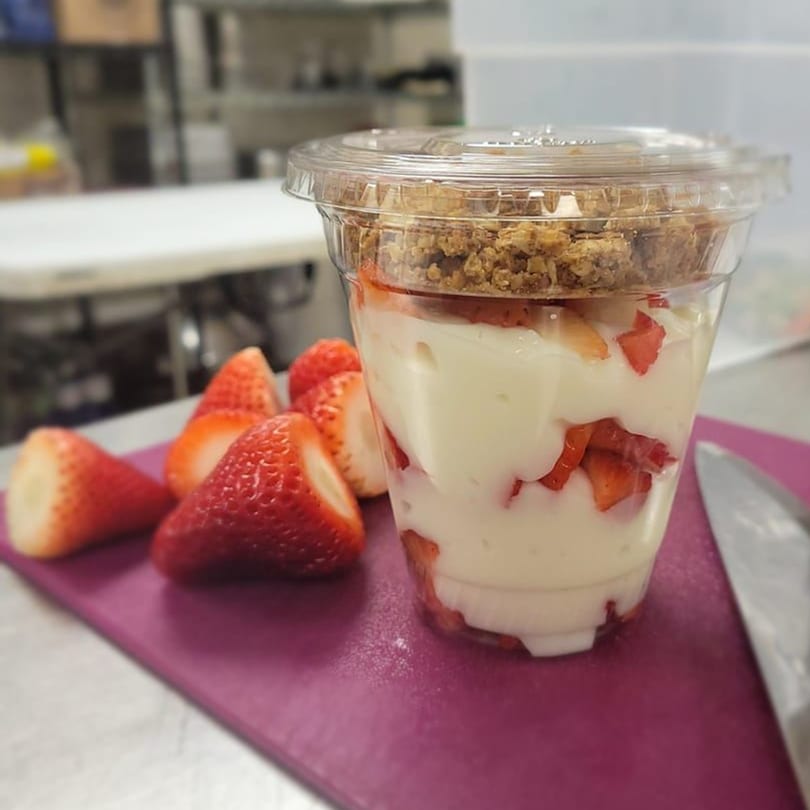 A clear plastic cup of yogurt parfait with strawberries and granola, next to fresh strawberries.