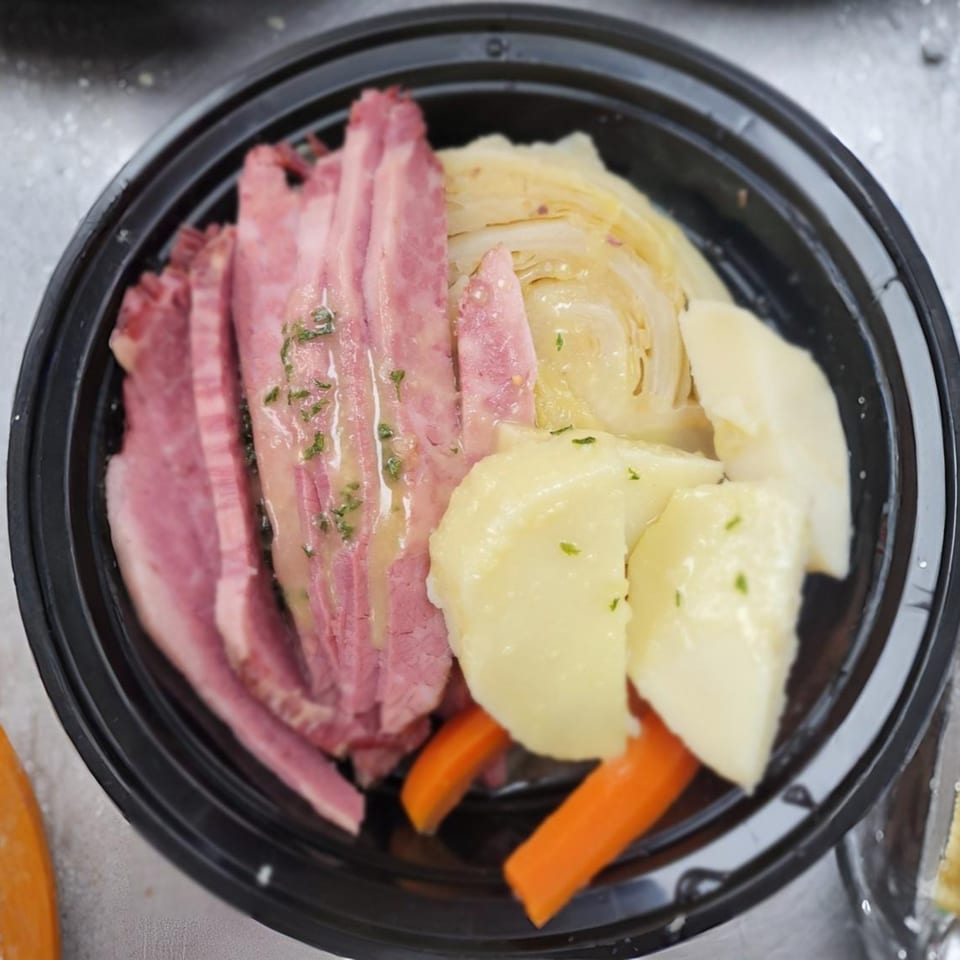 Corned beef with cabbage, potatoes, and carrots in a black takeout container.