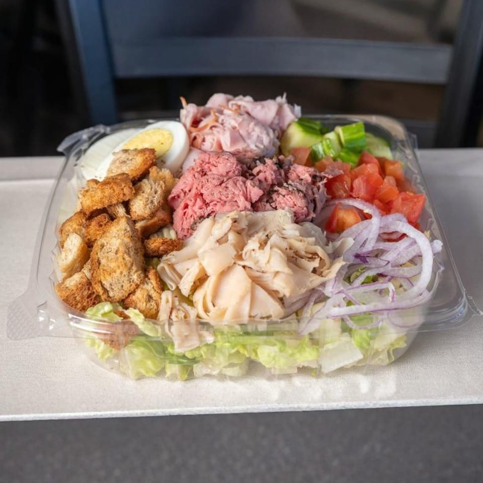 A clear plastic container filled with a chef salad, including meats, egg, croutons, and vegetables.