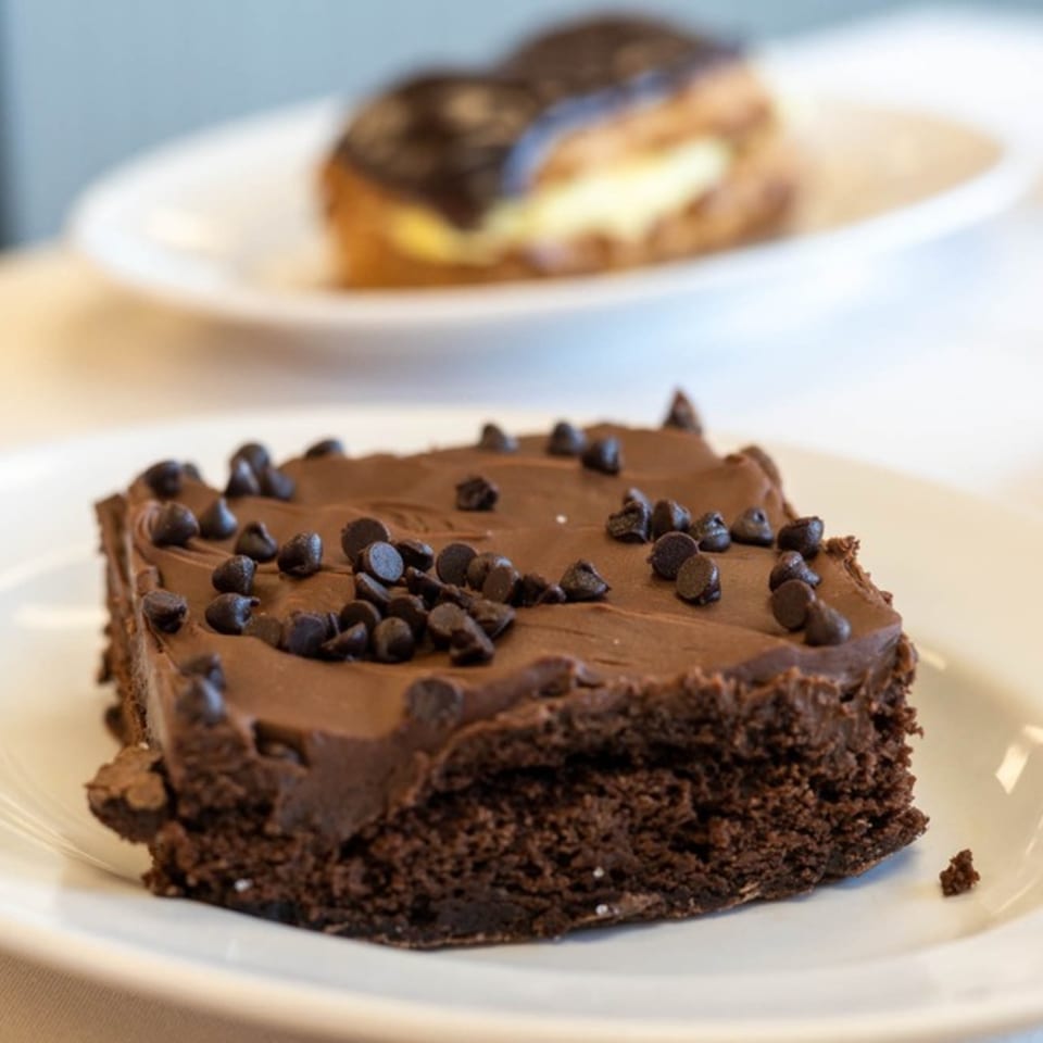A rich chocolate brownie with frosting and chocolate chips on a white plate, with a pastry in the background.