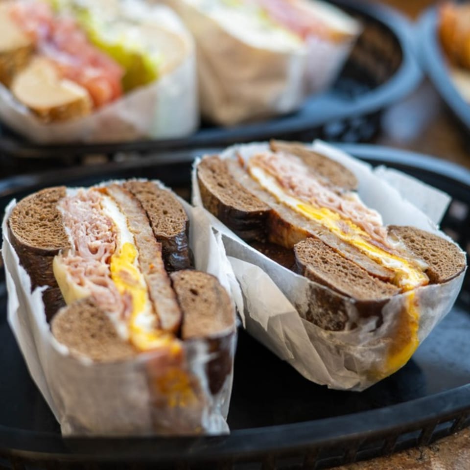 Two dark rye bagels with ham and egg, wrapped in paper, in a black basket.
